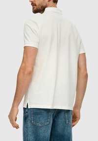 Mann in einem schlichten weißen Kurzarm-Polohemd und blauen Jeans, von hinten vor einem grauen Hintergrund gezeigt.