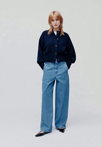 Jovem mulher com cabelo vermelho veste um cardigã azul escuro com botões, jeans largos de cor azul clara e sapatos pretos de bico contra um fundo simples.