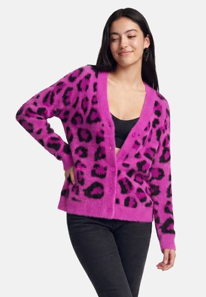 Junge Frau trägt eine leuchtend pinke, flauschige Strickjacke mit schwarzen Leopardenflecken, schwarzes Top und schwarze Hose, posiert mit einer Hand in der Hüfte.