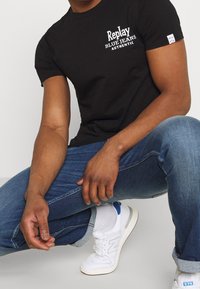 Czarny t-shirt z krótkim rękawem z białym napisem "Replay Blue Jeans Authentic", sparowany z niebieskimi dżinsami i białymi sneakersami New Balance.