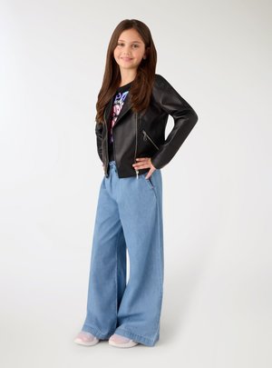 Ragazza giovane in piedi, indossa una giacca di pelle nera, una t-shirt grafica, jeans blu a gamba larga e scarpe di colore chiaro su uno sfondo semplice.