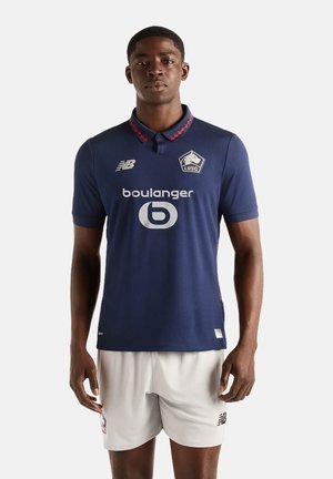 Modello maschile che indossa una maglia da calcio LOSC blu navy con loghi bianchi e pantaloncini grigio chiaro, in piedi davanti a uno sfondo semplice.