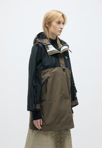 Brūna un melna ūdensizturīga jaka ar rāvējslēdzēja priekšpusi, kapuci un regulējamiem piedurkņiem. Iekļauj rāvējslēdzēja kabatas un gludu tekstūru.