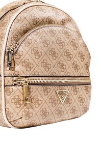 Mochila beige con un patrón y forma redondeada, que cuenta con cremalleras en tono dorado, un bolsillo frontal y una placa con el logo de Guess. Textura de tela suave.