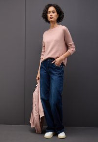 Maglione rosa chiaro, jeans a gamba larga blu, in mano una giacca rosa chiaro. Sneakers bianche con suole texture. Sfondo neutro.