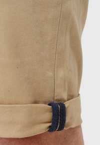 Beige stofshorts med en rullet kant, udstyret med en mørkeblå stofdetalje der er hyggeligt indpakket omkring afslutningen. Teksturen ser glat og holdbar ud.