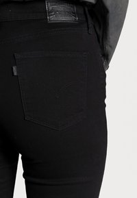 Black denim jeans featuring a distinctive back pocket design with curved stitching. The waistband includes a black leather label.