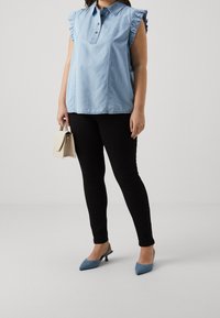 Top in denim azzurro con maniche arricciate, pantaloni neri aderenti e décolleté blu con cinturino. La modella tiene una borsa beige.