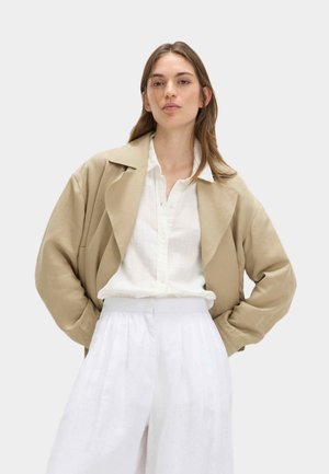 Vrouw draagt een beige oversized blazer over een wit overhemd met knopen en witte, losse, hooggetailleerde broek, staand met handen in de zakken.