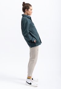 Maglietta a manica lunga color verde scuro con due tasche frontali e un logo verde, abbinata a pantaloni tapered beige chiaro e sneakers bianche con dettagli neri.