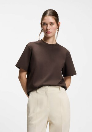 Donna con una maglietta a maniche corte marrone scuro infilata in pantaloni beige a vita alta, in piedi con le mani dietro la schiena su sfondo bianco.