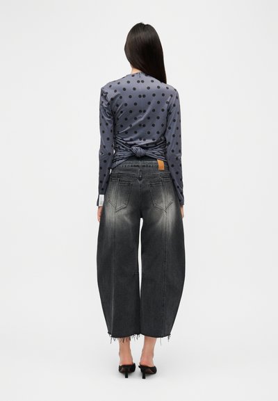 Γκρι τοπ με μακριά μανίκια και πουά σχέδιο, με λεπτομέρεια κόμβου στην πλάτη, συνδυασμένο με ψηλόμεσα, φαρδιά μαύρα τζιν παντελόνια με ξεθωριασμένο μπροστινό μέρος και φθαρμένο τελείωμα.