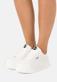 Bílé plátěné tenisky s tlustou, texturovanou podešví. Mezi vlastnosti patří kulatý černý nášivka s logem a tradiční šněrovací design.