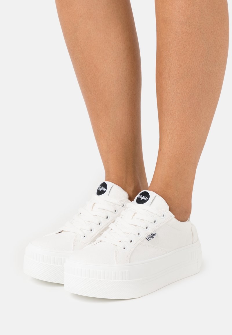 Bílé plátěné tenisky s tlustou, texturovanou podešví. Mezi vlastnosti patří kulatý černý nášivka s logem a tradiční šněrovací design.