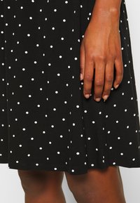 Robe noire à pois blancs, en tissu de coton, avec une longueur au genou, présentant des accents de piqûre visibles près de l'ourlet et sur le côté.
