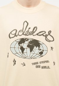 Μπεζ πουκάμισο με στυλιζαρισμένη υδρόγειο, αστέρια, το κείμενο «Three Stripes. One World.» και ένα μεγάλο καλλιγραφικό λογότυπο πάνω από την υδρόγειο.