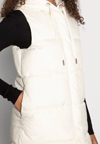 Weste in Creme mit Kapuze, Reißverschluss vorne, Kordelzug und gestepptem Design. Schwarzes Langarmshirt, das darunter hervorschaut.