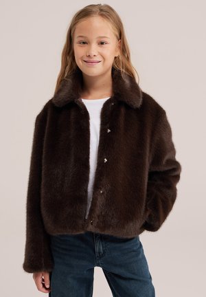 Une jeune fille souriante porte une veste brun foncé à la texture de fourrure par-dessus une chemise blanche et un jean bleu, devant un fond uni.