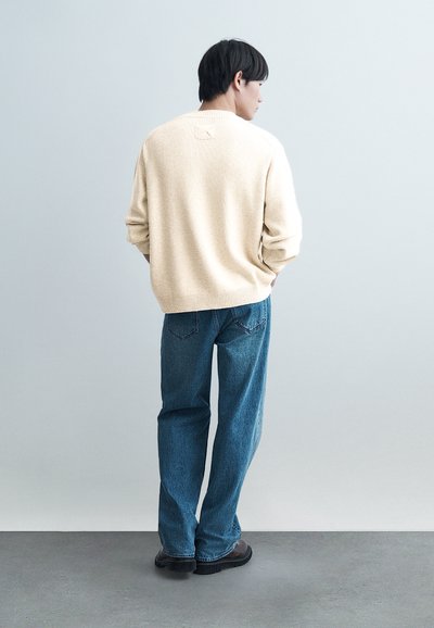 Homme debout, dos tourné vers la caméra, portant un pull crème, un jean bleu et des chaussures noires, sur un fond gris clair uni.