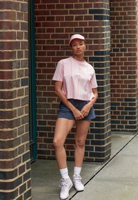 Kortärmad rosa randig skjorta, tillsammans med marinblå shorts. Bär vita sneakers och en matchande keps, står mot tegelpelare.