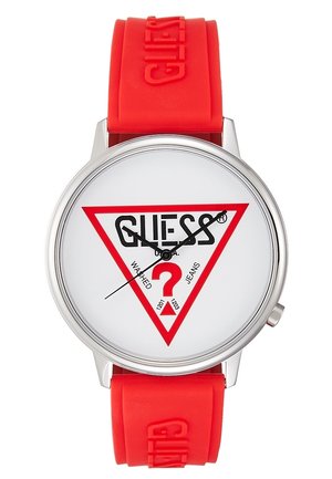 Okrągły srebrny zegarek z białą tarczą z czerwonym trójkątem i czarnym logo GUESS, czerwonym znakiem zapytania oraz czerwonym silikonowym paskiem z wytłoczonym logo GUESS.