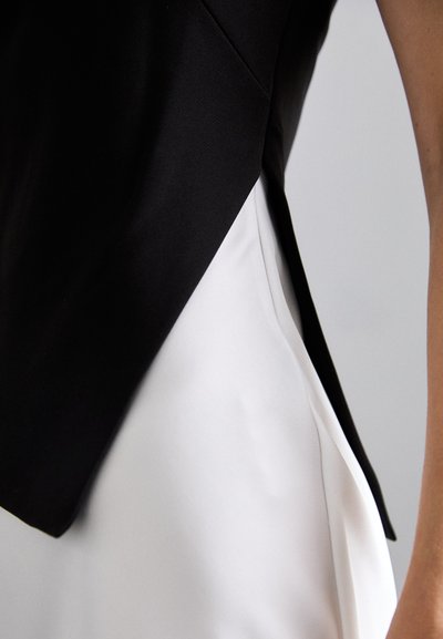 Black sleeveless top with an angled hem paired with a white bottom. Smooth, lightweight fabric, featuring contrasting colors and clean lines.
