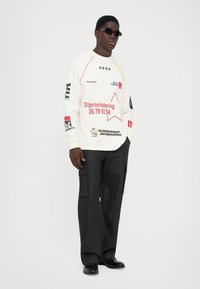 Man wearing black sunglasses, white long-sleeve shirt with red and black text and symbols, black cargo pants, and black shoes, standing against white background.
