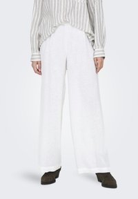 Pantalons larges en lin blanc, avec une finition texturée, associés à une chemise à rayures ample dans des tons neutres. Des chaussures épaisses et sombres complètent le look.