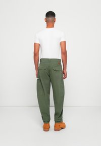 Man facing away wearing white short-sleeve shirt, green cargo pants with back pockets, and tan work boots against a plain white background.