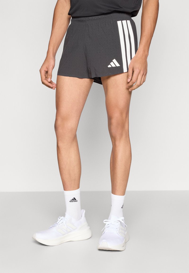 Shorts de sport noirs à texture perforée, avec trois bandes blanches et logo sur le côté. Portés avec des chaussures et des chaussettes de sport blanches.