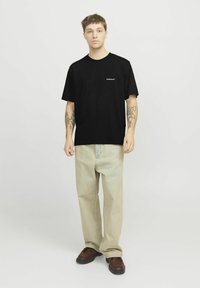 Sort bomulds t-shirt med korte ærmer og et lille hvidt logo på brystet, parret med beige brede bukser og brune sko.
