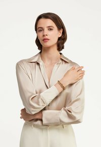 Cămașă din satin taupe cu mâneci lungi, cu manșete cu nasturi, având un guler lejer. Brățară din lanț de aur pe încheietura mâinii, textură moale și netedă.
