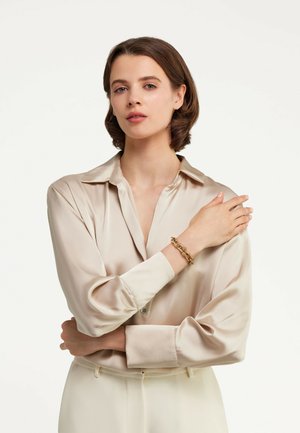 Long-sleeve, taupe satin shirt with buttoned cuffs, featuring a relaxed collar. Gold chain bracelet on the wrist, soft, smooth texture.
