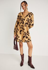 Φόρεμα κοντό μανίκι, μέχρι το γόνατο, σε χρυσό σατέν με μαύρο λεοπάρ σχέδιο, μαζεμένο στη μέση, φορεμένο με μποτάκια μέχρι το γόνατο σε μπορντό και τσάντα σε αντίστοιχο χρώμα.