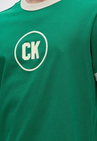 Πράσινο T-shirt με στρογγυλή λαιμόκοψη, λευκό γιακά και τα αρχικά "CK" μέσα σε έναν λευκό κύκλο στο στήθος.