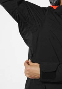 Černá bunda se síťovou ventilací v podpaží na zip, nastavitelnými manžetami na suchý zip a aerodynamickým designem. Obsahuje detailní záběr ruky, která upravuje zip.