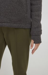 Sivá štruktúrovaná bunda na zips s otvormi na palce, skombinovaná s olivovozelenými priliehavými nohavicami. Detailný záber na rukávy a ruku na páse.