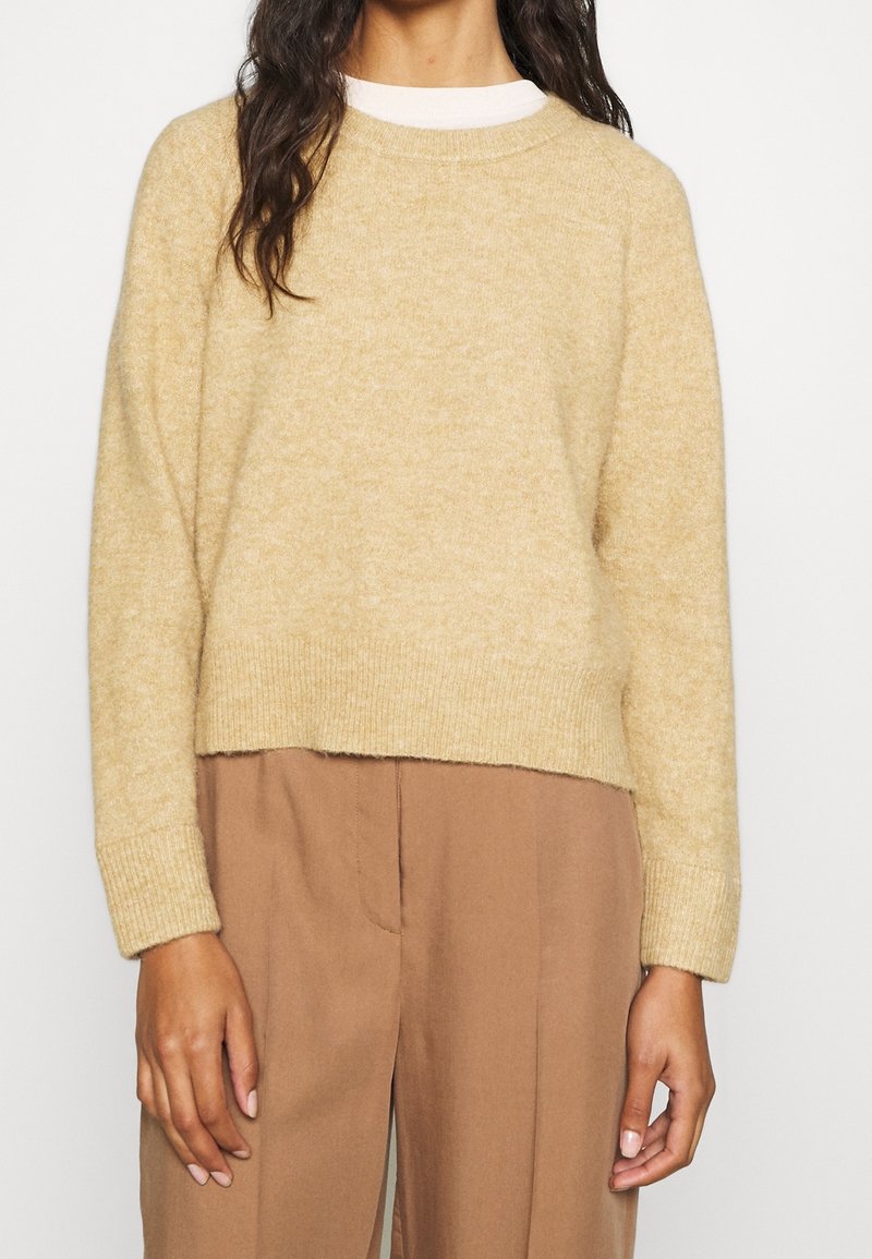 Femme portant un pull beige duveteux à manches longues sur une chemise blanche, associé à un pantalon marron clair taille haute, debout devant un fond uni.
