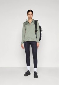 Kvinne som står iført en grønn zip-up jakke, svarte leggings, svarte joggesko, hvite sokker, og en svart ryggsekk på den ene skulderen.