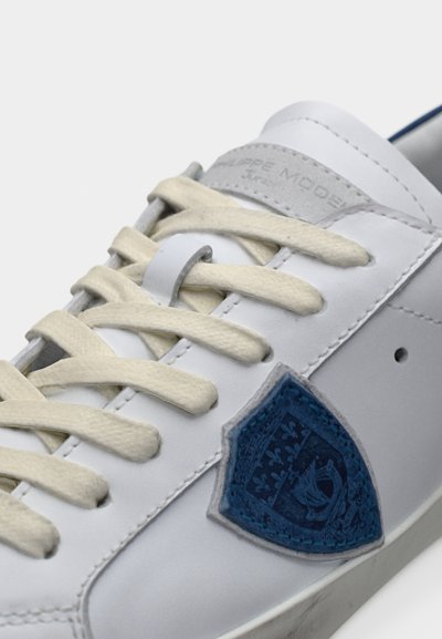 Λευκό αθλητικό παπούτσι με κρέμ κορδόνια, μπλε έμβλημα ασπίδας στο πλάι και γκρι ετικέτα στη γλώσσα με ραμμένη επωνυμία.