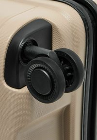 Close-up van een beige, gestructureerde kofferhoek met dubbele zwarte draaide wielen voor 360-graden mobiliteit.
