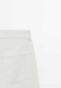 Vue arrière d'un jean en denim blanc montrant la ceinture, la boucle de ceinture et une seule poche arrière sur un fond clair uni.