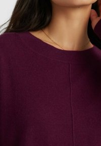 Burgunderfarbener Strickpullover mit weitem Rundhalsausschnitt und sichtbaren Nahtdetails, kombiniert mit einer zarten goldenen Halskette.