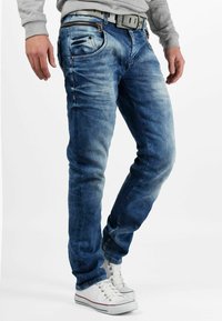 Jean en denim de lavage bleu clair avec une coupe slim, présentant une fermeture éclair, cinq poches et des détails de couture. Associé à des baskets en toile blanches.