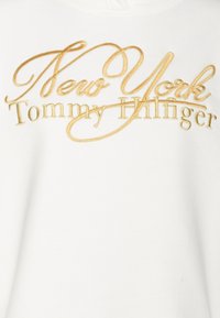 Tessuto bianco con testo ricamato in oro che recita "New York" in corsivo sopra "Tommy Hilfiger" in carattere serif maiuscolo.