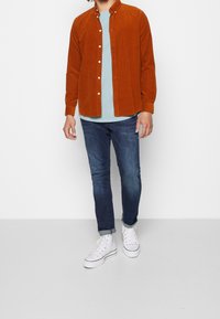 Rostfärgad kordflosshirt över en ljusblå T-shirt; matchad med mörkblå jeans och vita sneakers. Knapparna på skjortan är synliga.