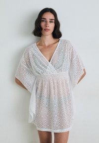 White lace dress with a V-neck, short sleeves, and an elastic waist. Features intricate oval patterns throughout. Lightweight and sheer fabric.