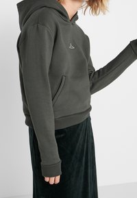 Tmavě zelená mikina vyrobená z měkké tkaniny, s přední kapsou a žebrovanými manžetami. Na hrudi se nachází malé bílé logo ramínka.