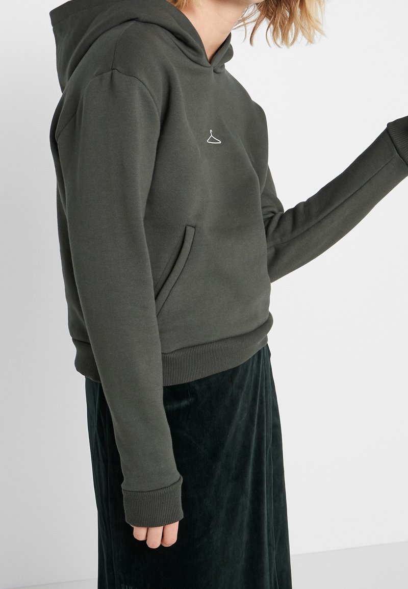 Tmavě zelená mikina vyrobená z měkké tkaniny, s přední kapsou a žebrovanými manžetami. Na hrudi se nachází malé bílé logo ramínka.