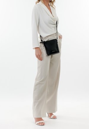 Musta nahkainen olkalaukku, jossa säädettävät hihnat. Pehmeä pinta, minimalistinen muotoilu. Malli pukeutuneena vaaleaan toppiin ja housuihin, seisoo.